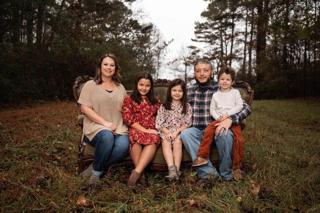 family photo of husband and wife and three children sitting on a couch in a clearing
