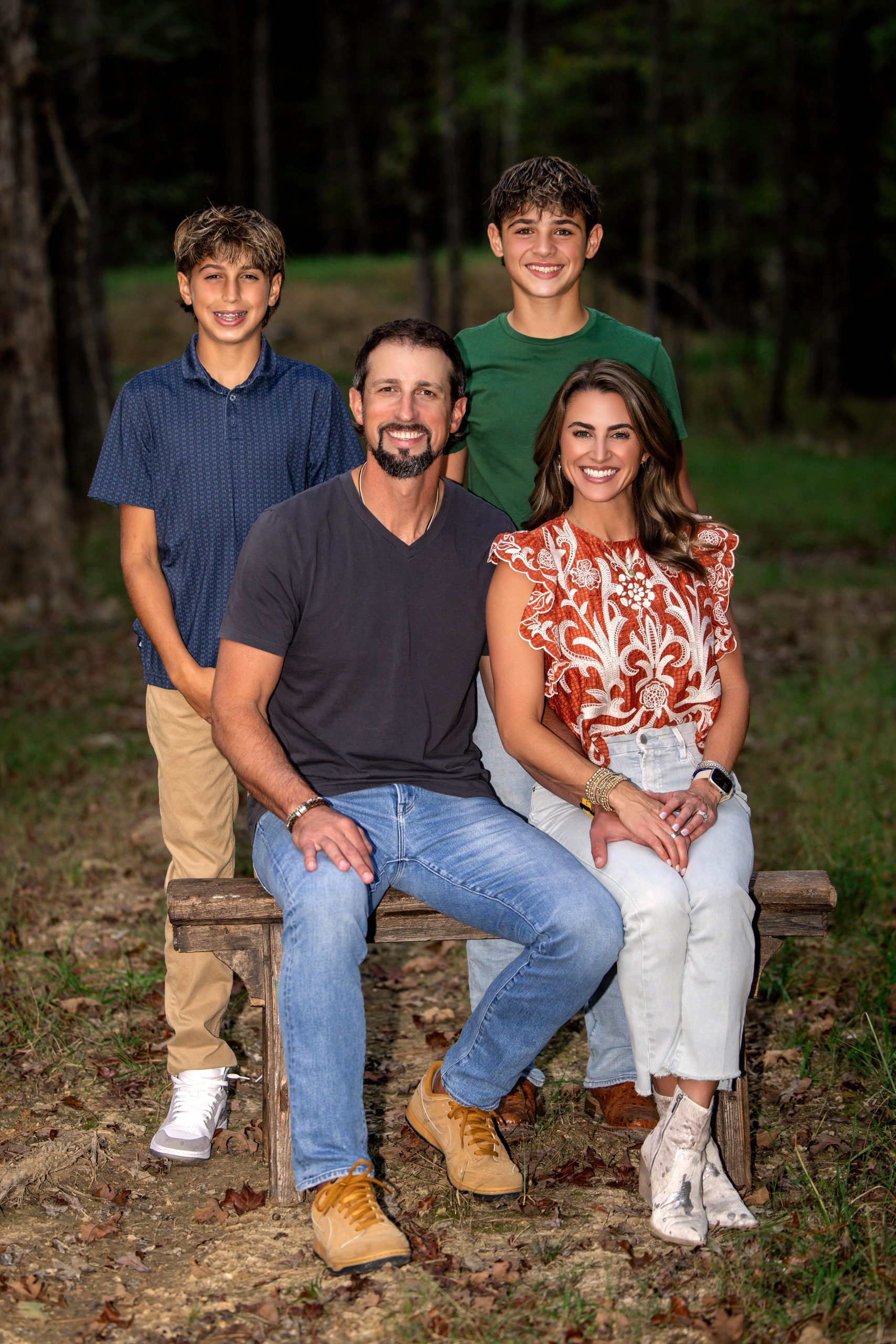 Brodie and Kelli Croyle sit on a wooden bench among trees, with Luke and Sawyer standing behind them.