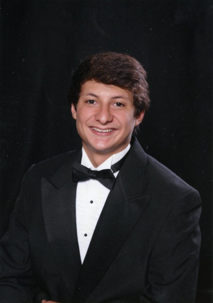 Gilbert smiles in a black tuxedo, white shirt and bow tie, posed against a dark backdrop.