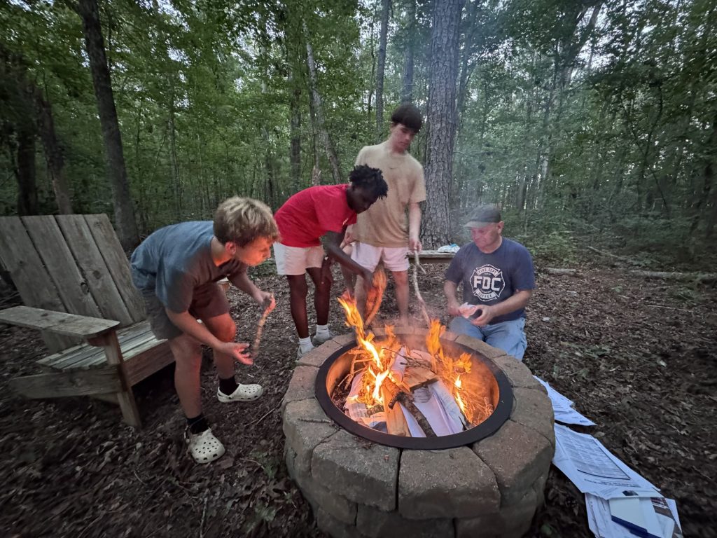 Boys' Ranch boys and a man circle a fire pit in the woods at dusk.