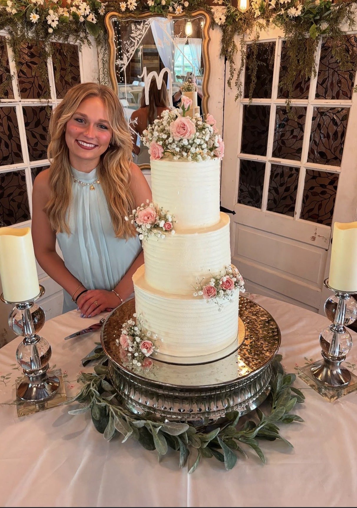 Taylor smiles beside a tall white tiered cake decorated with pink roses.