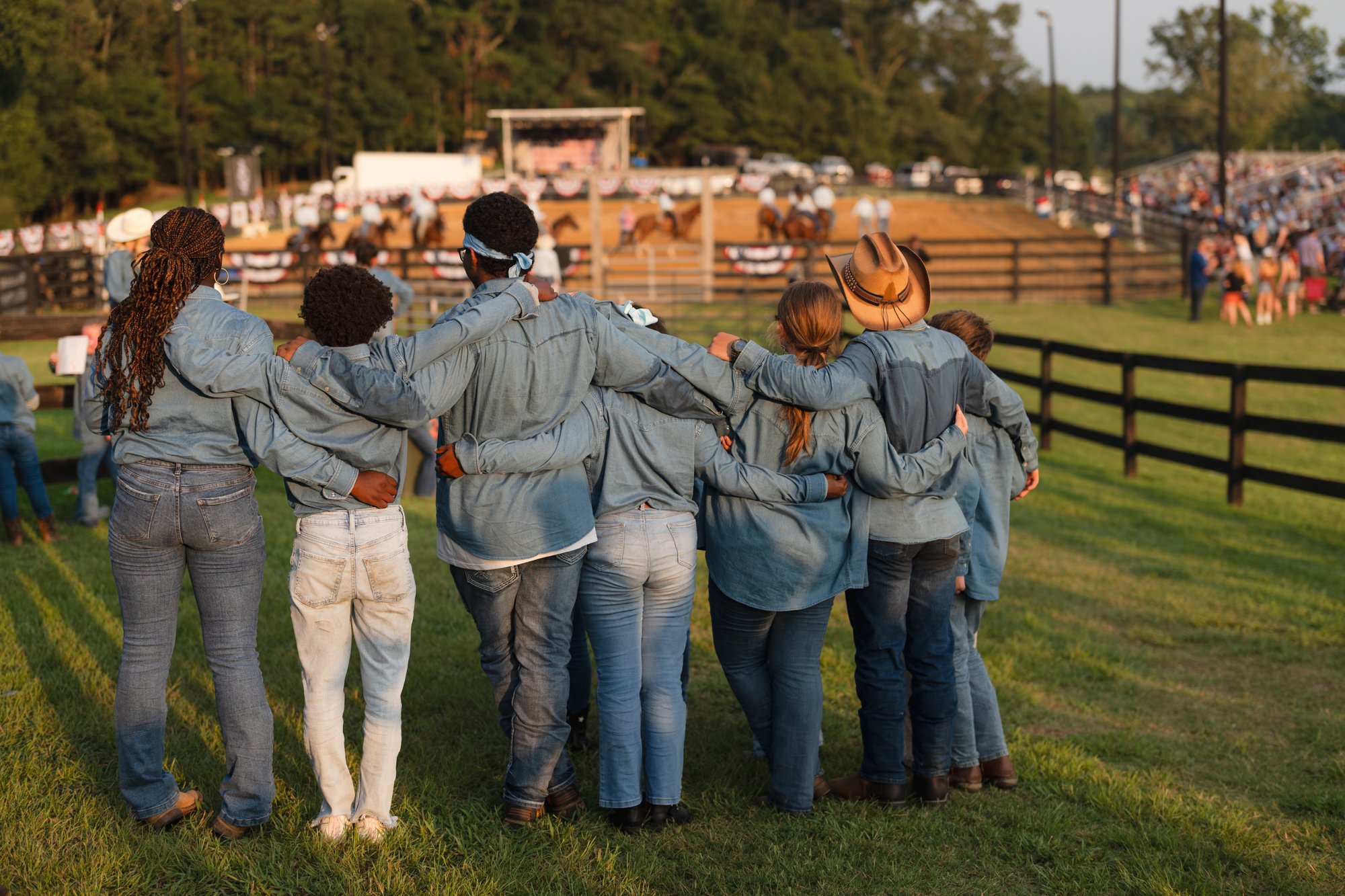 A group in matching denim shirts stands arm in arm, backs to the camera, watching riders in a dirt arena.