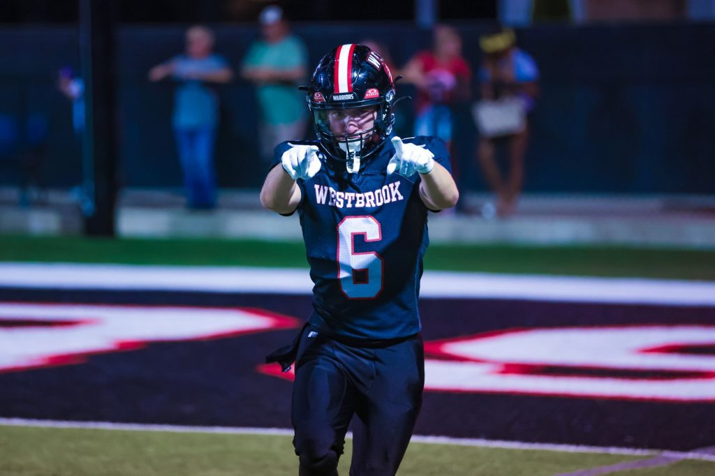 A Westbrook football player in helmet and gloves points both hands toward the camera under the lights.