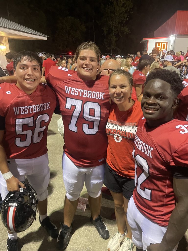 Three Westbrook football players and a smiling woman crowd together for a photo at night.