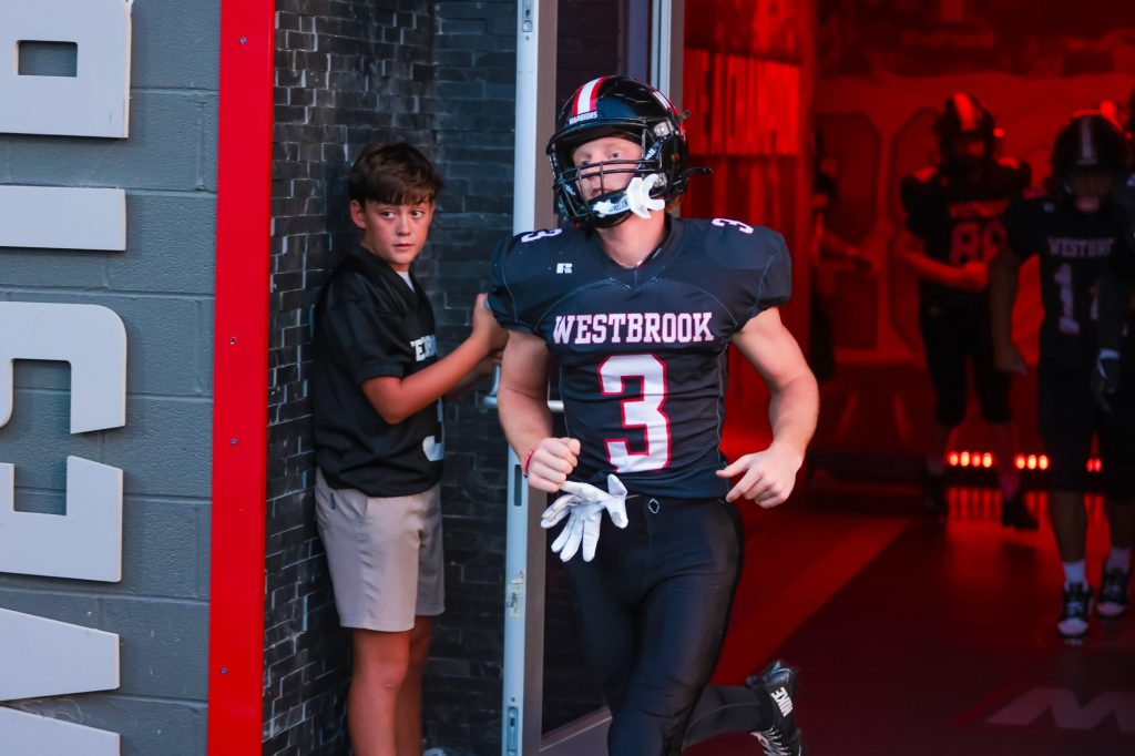 A Westbrook football player in full uniform steps out of a doorway as a boy looks on.