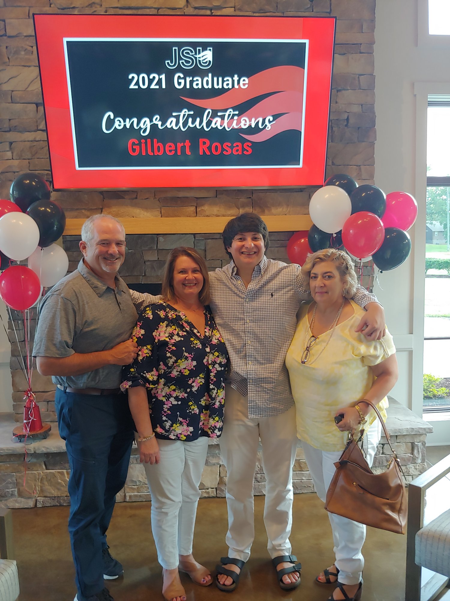 Gilbert celebrates his college graduation with three adults beneath a congratulations banner and balloons.