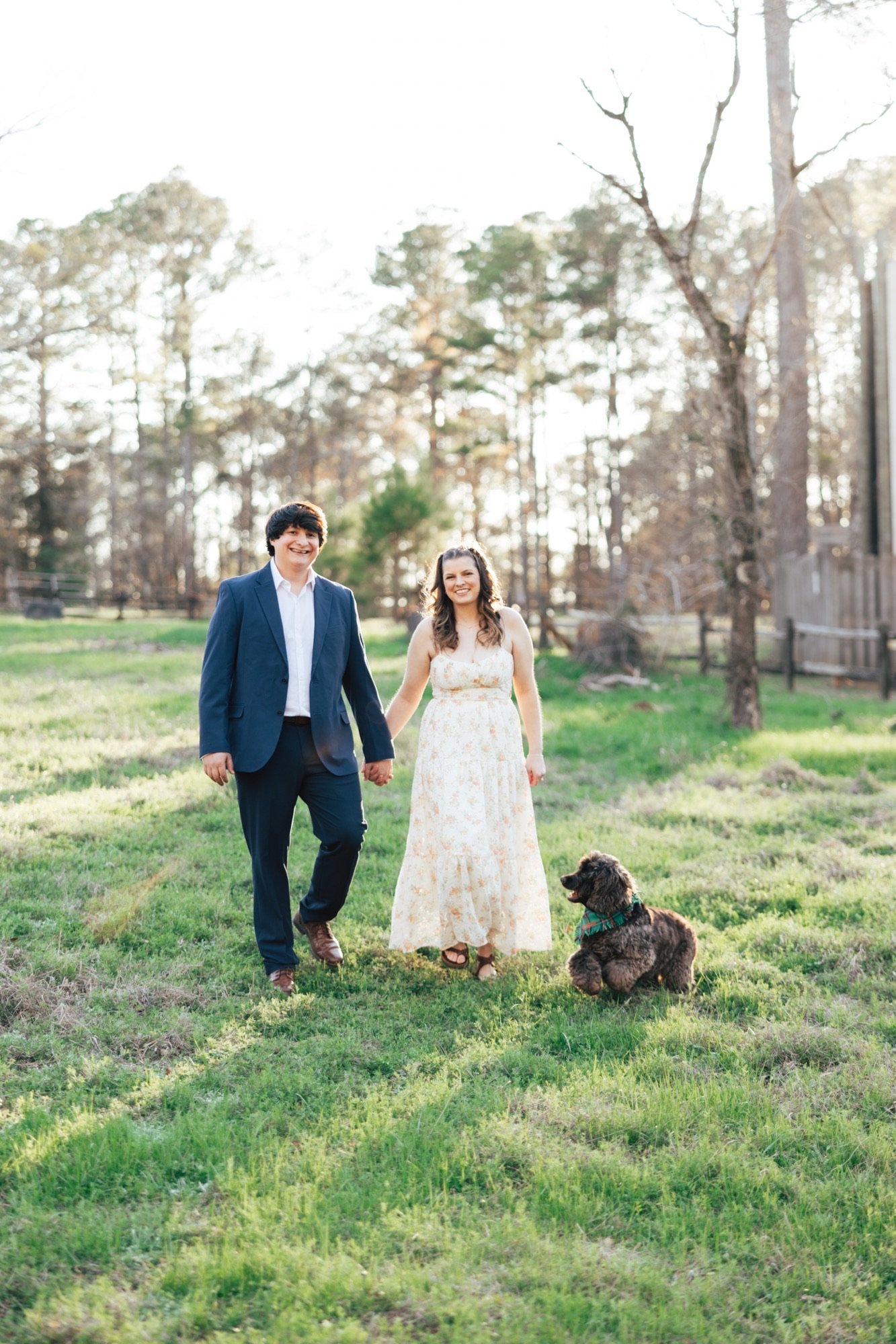 Gilbert and his fiancée, Kelsey, walk hand in hand across a grassy field with a dog beside them.