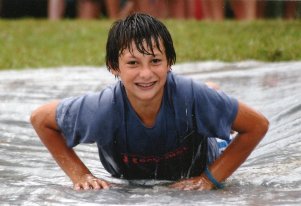 A soaking-wet Gilbert as a boy, smiling up from a plastic water slide stretched across the lawn.