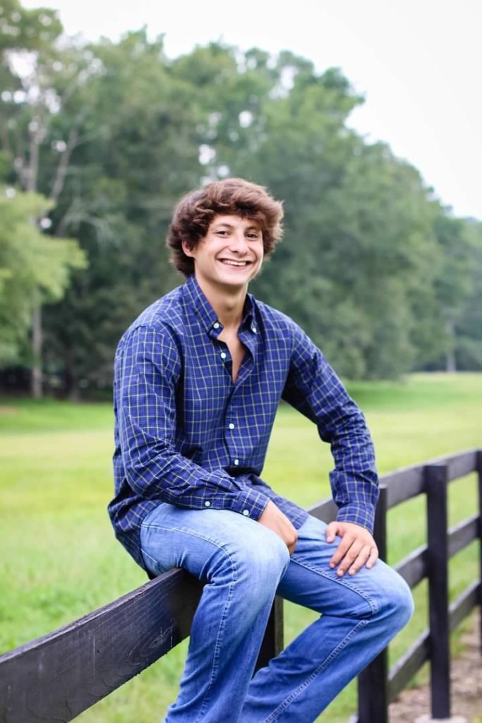 Gilbert, smiling in a blue plaid shirt and jeans, sits on a black fence with tall trees behind him.