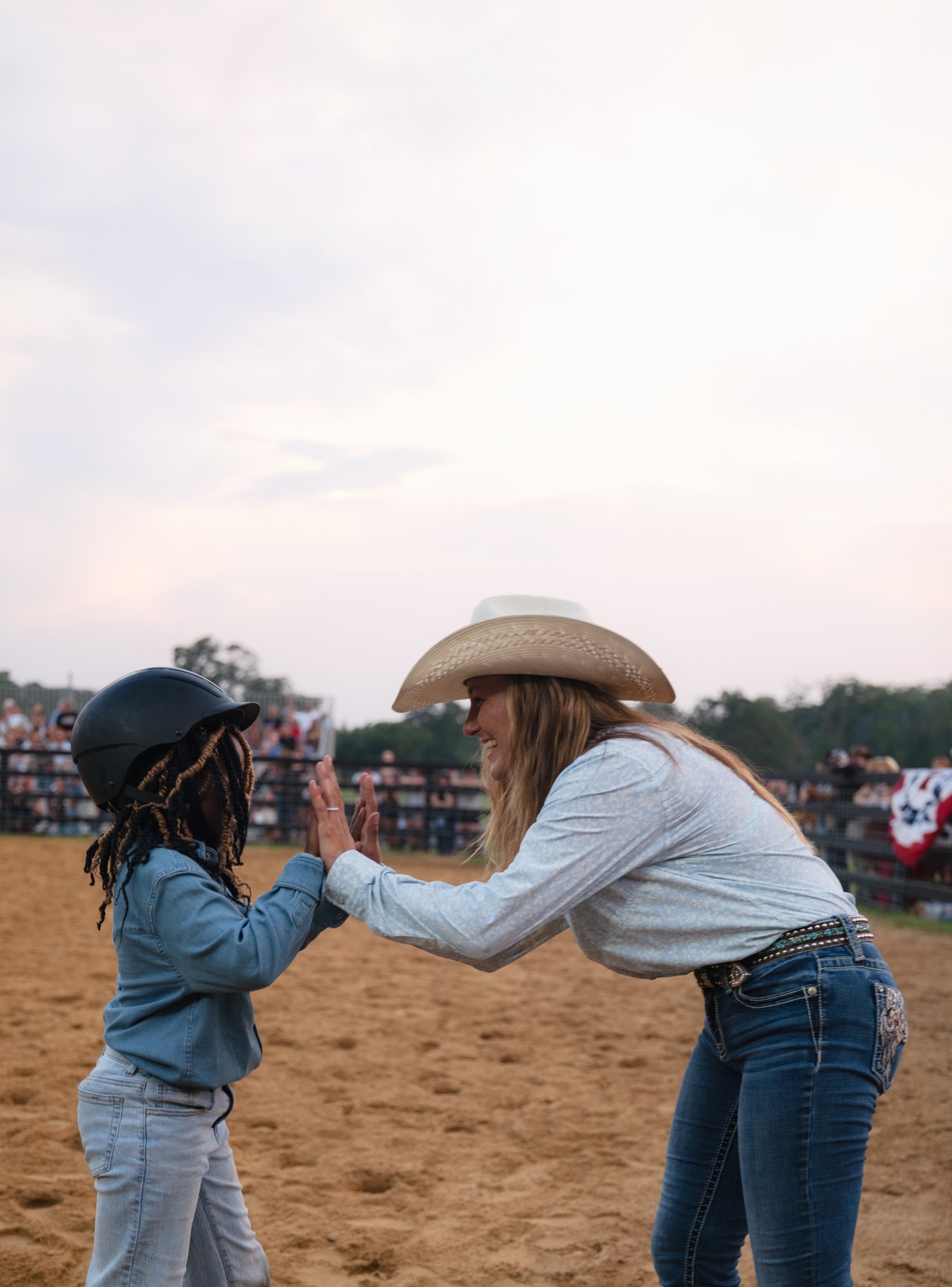A child in a helmet high-fives a smiling woman in a cowboy hat in a dirt arena as spectators watch.