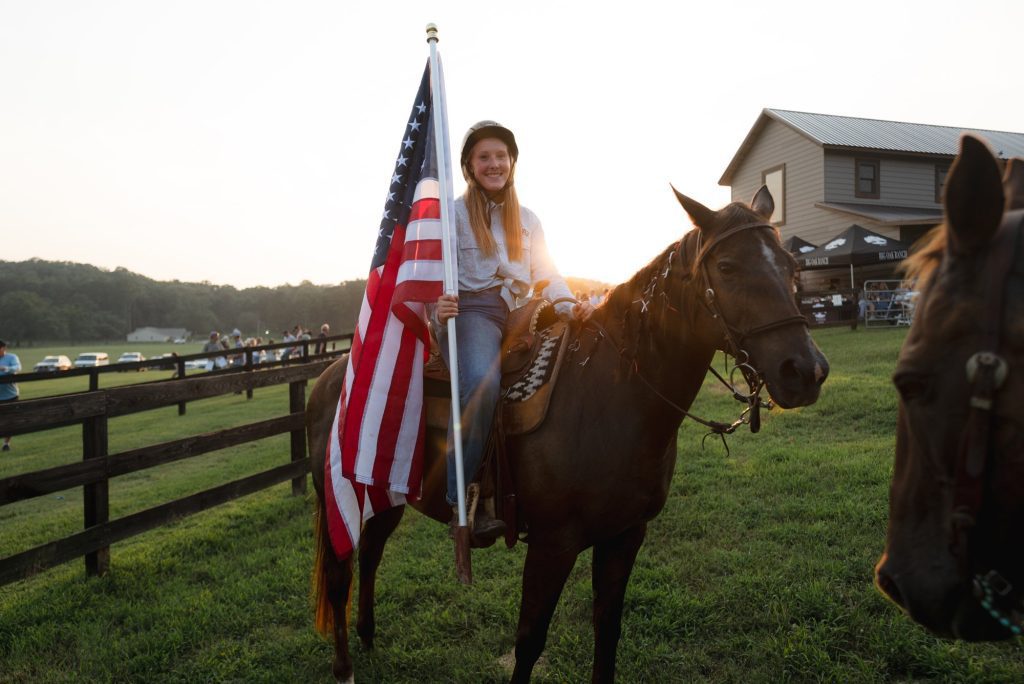 Makenzie, Girls' Ranch Head Wrangler, smiles on horseback holding an American flag at sunset.
