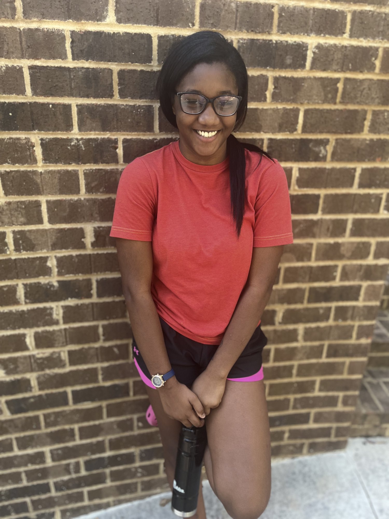 Yanna at 16 years old, smiling in glasses and a red shirt in front of a brick wall.