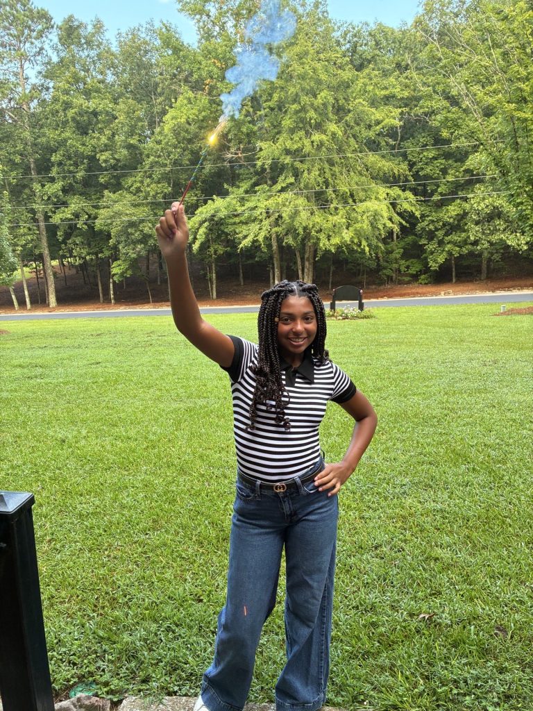 A Girls' Ranch girl in a striped shirt smiles and waves on a green lawn.