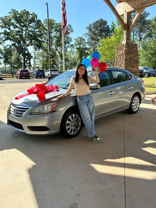 A young woman leans against a silver car decorated with a red bow and balloons.
